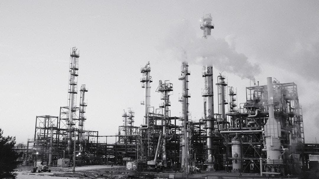 Oil refinery with distillation towers and smoking stacks under a cloudy sky, illustrating the scale of emissions requiring fenceline monitoring
